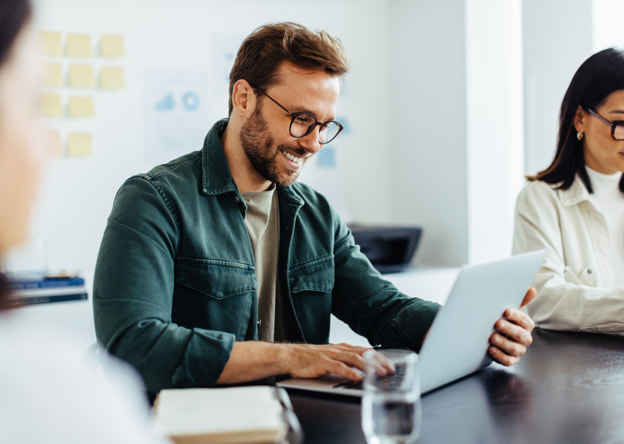 Smiling man working at a laptop while in a meeting room with coworkers.