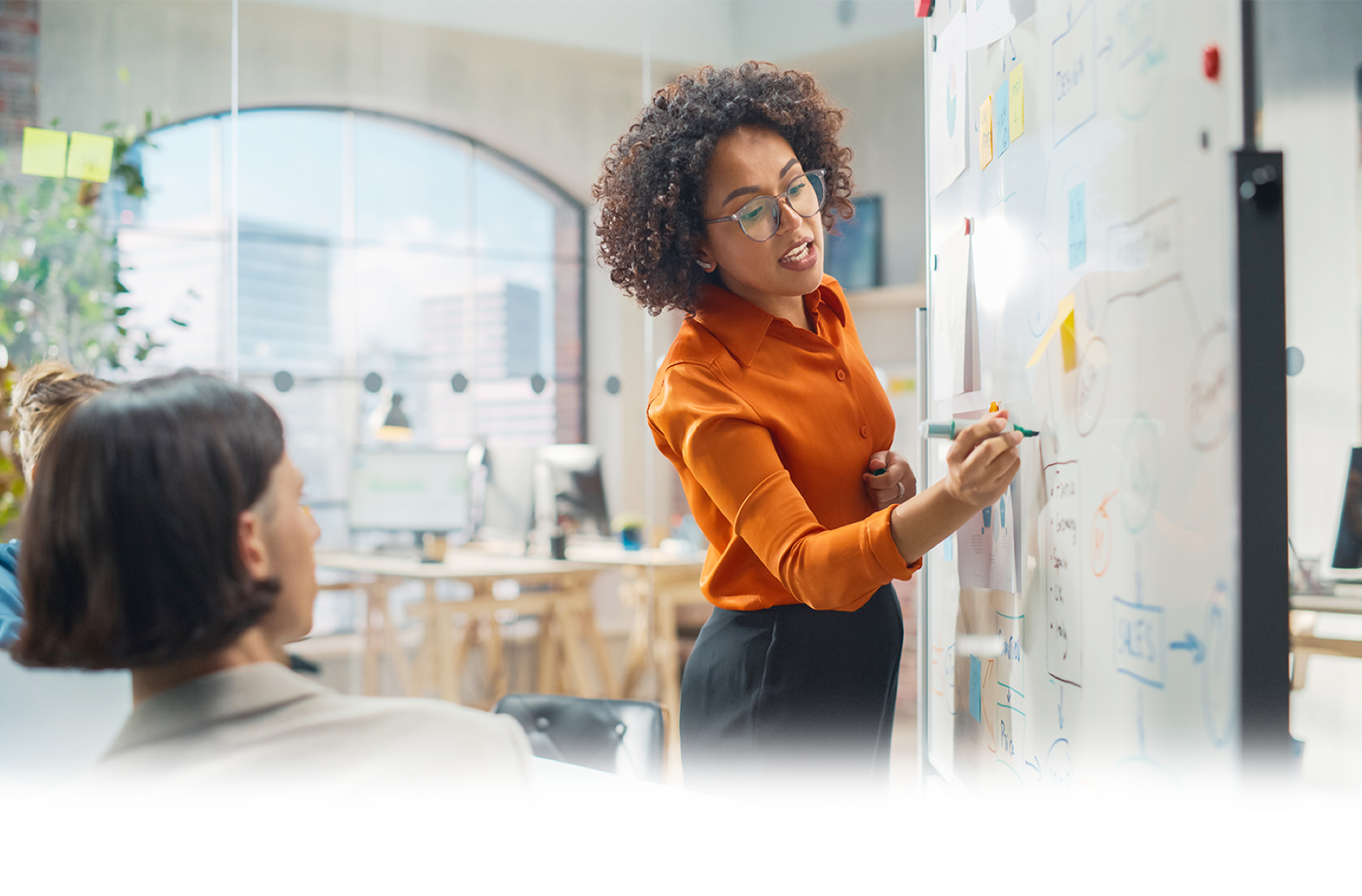 Woman in a meeting writing on a colorful whiteboard in a bright office setting with an orange ‘Challenge the Process’ banner at the bottom.