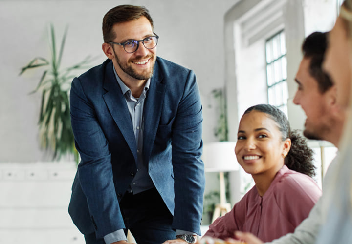 Man leaning over a desk, presenting information to colleagues in a collaborative office environment.