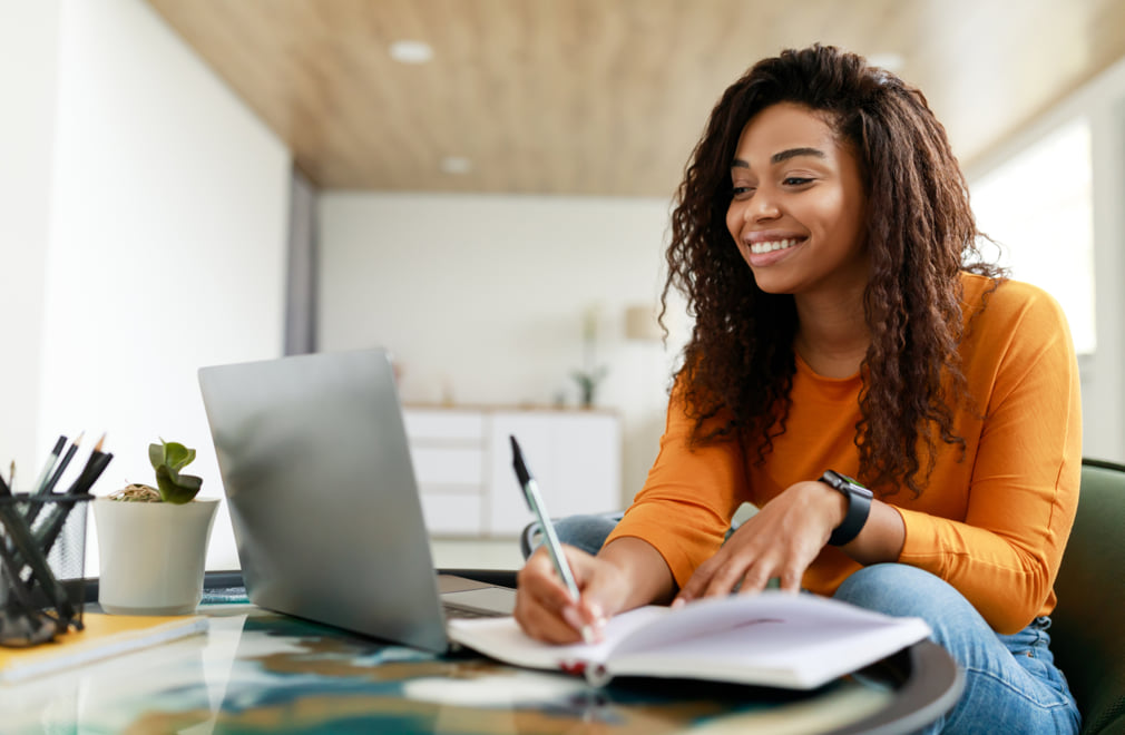 Young female student writing in a notebook while looking at a laptop.
