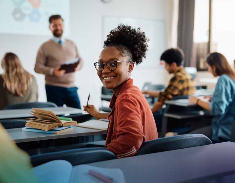 Smiling woman sitting at a desk in a classroom setting with a teacher presenting something in the background.