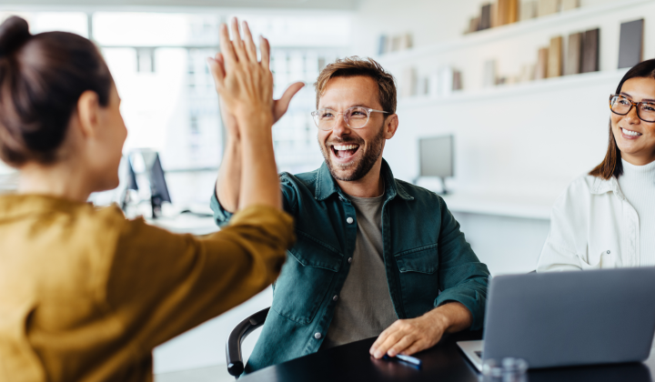 Two coworkers high fiving while working in front of a laptop.